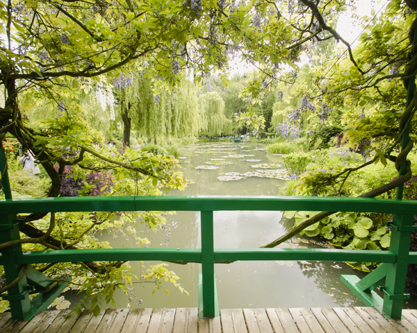 A picturesque bridge with green railings crosses a tranquil pond, framed by vibrant trees in Monet's Giverny garden.