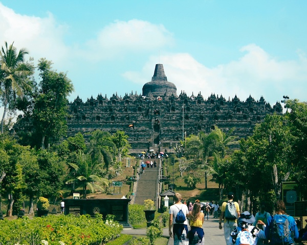 A group of people walks along a pathway, admiring the iconic Borobudur Temple in Indonesia's beautiful landscape.