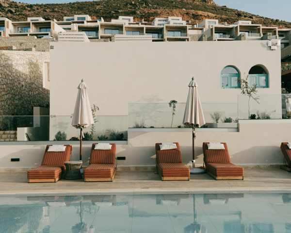 A blue pool in Greece, featuring lounge chairs and umbrellas, set against a mountainous backdrop.