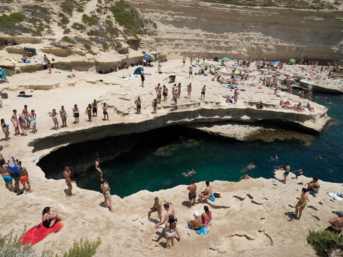 A swimming hole in Malta during the summer, crowded with people in the sun and crystal-clear waters, surrounded by rocky cliffs.