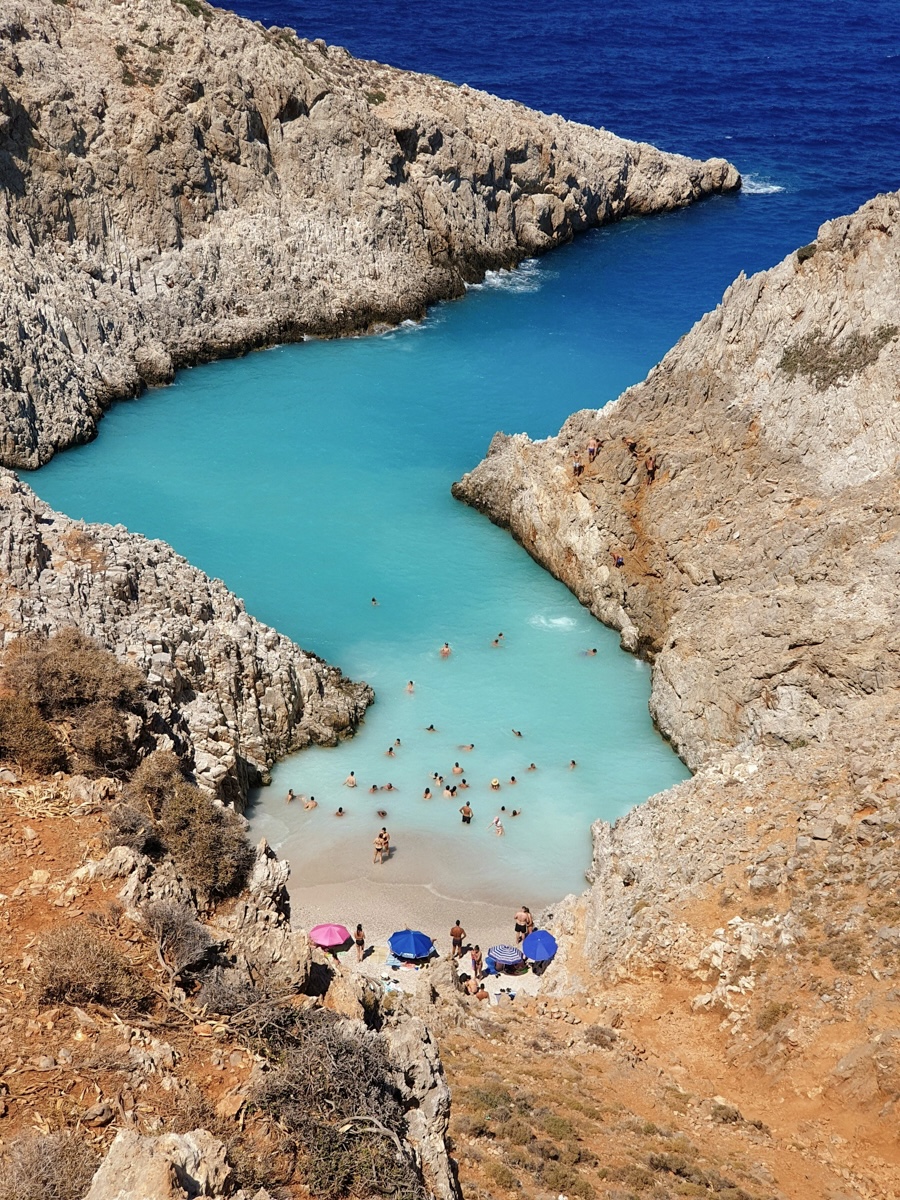 A beautiful beach in Crete, Greece, with golden sand, clear turquoise waters, and sunbathers relaxing under the warm sun, framed by rugged cliffs.