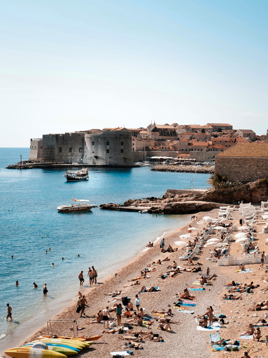 A beach in Dubrovnik, Croatia, featuring pebbly shores, crystal-clear waters, and historic city walls in the background, with people enjoying the summer day.