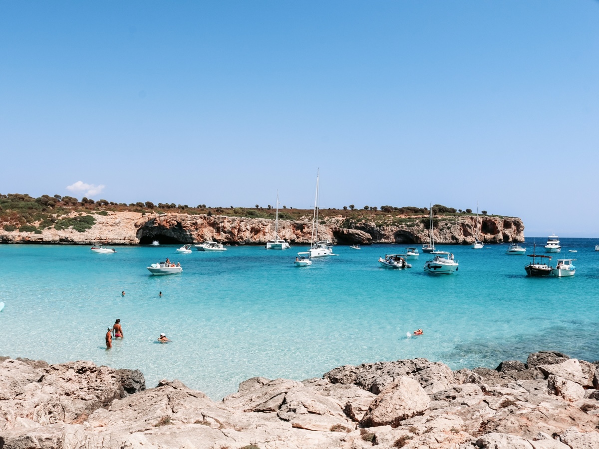 A beach in Mallorca, Spain, with soft white sand and azure waters, as beachgoers bask in the sun and swim in the sea.