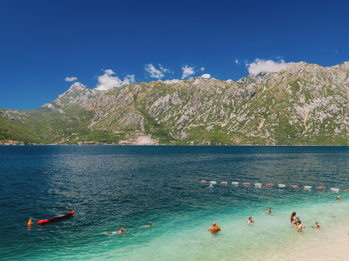 A tranquil beach in the Bay of Kotor, surrounded by mountains and calm, clear waters, with people enjoying the sun and water.