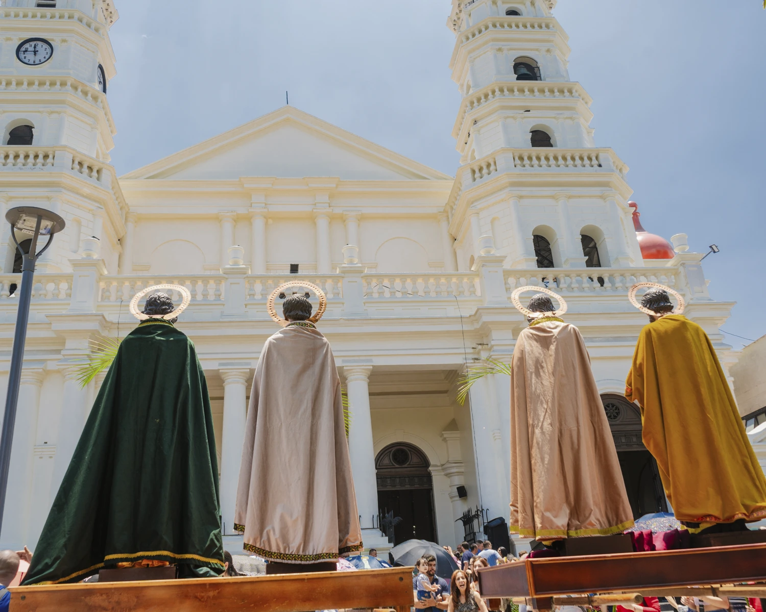 Three robed participants standing in front of a large white church during a Semana Santa procession, with a crowd gathered around.