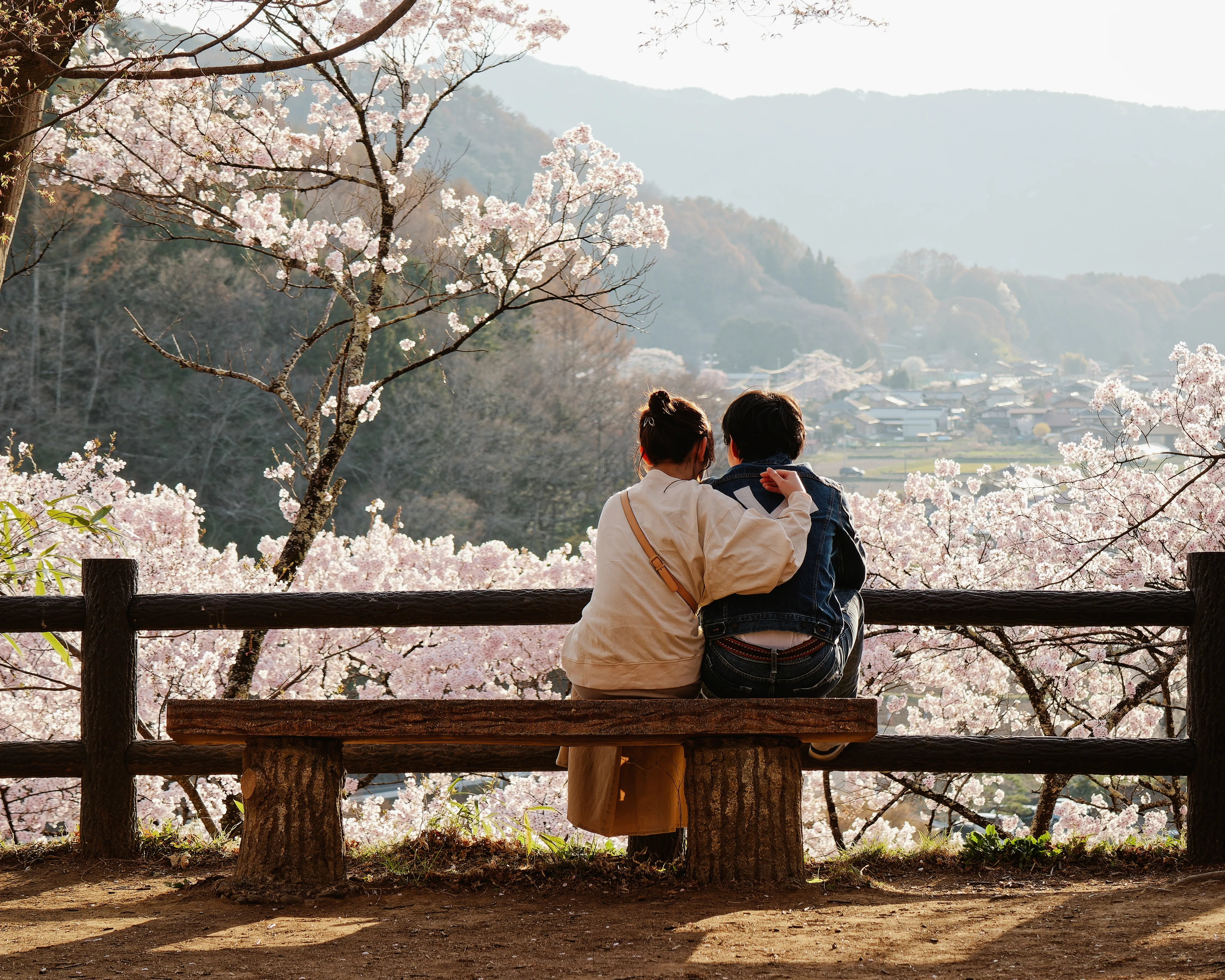 Two travellers sitting on a bench overlooking cherry blossoms and mountains in Japan during spring.