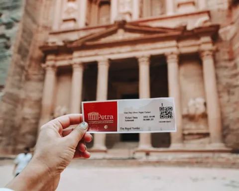 A hand holding out a ticket to the ancient rock-cut architecture of Petra, Jordan, with its iconic Treasury building carved into red sandstone cliffs