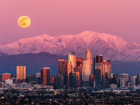 Los Angeles downtown skyline at sunset with a full moon rising over snow-capped San Gabriel Mountains — a direct flight destination from Australia