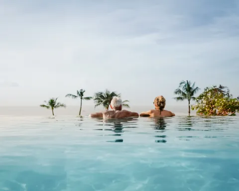 Older travellers relaxing in an infinity pool overlooking a tropical beach with palm trees, representing senior travel and holiday experiences.