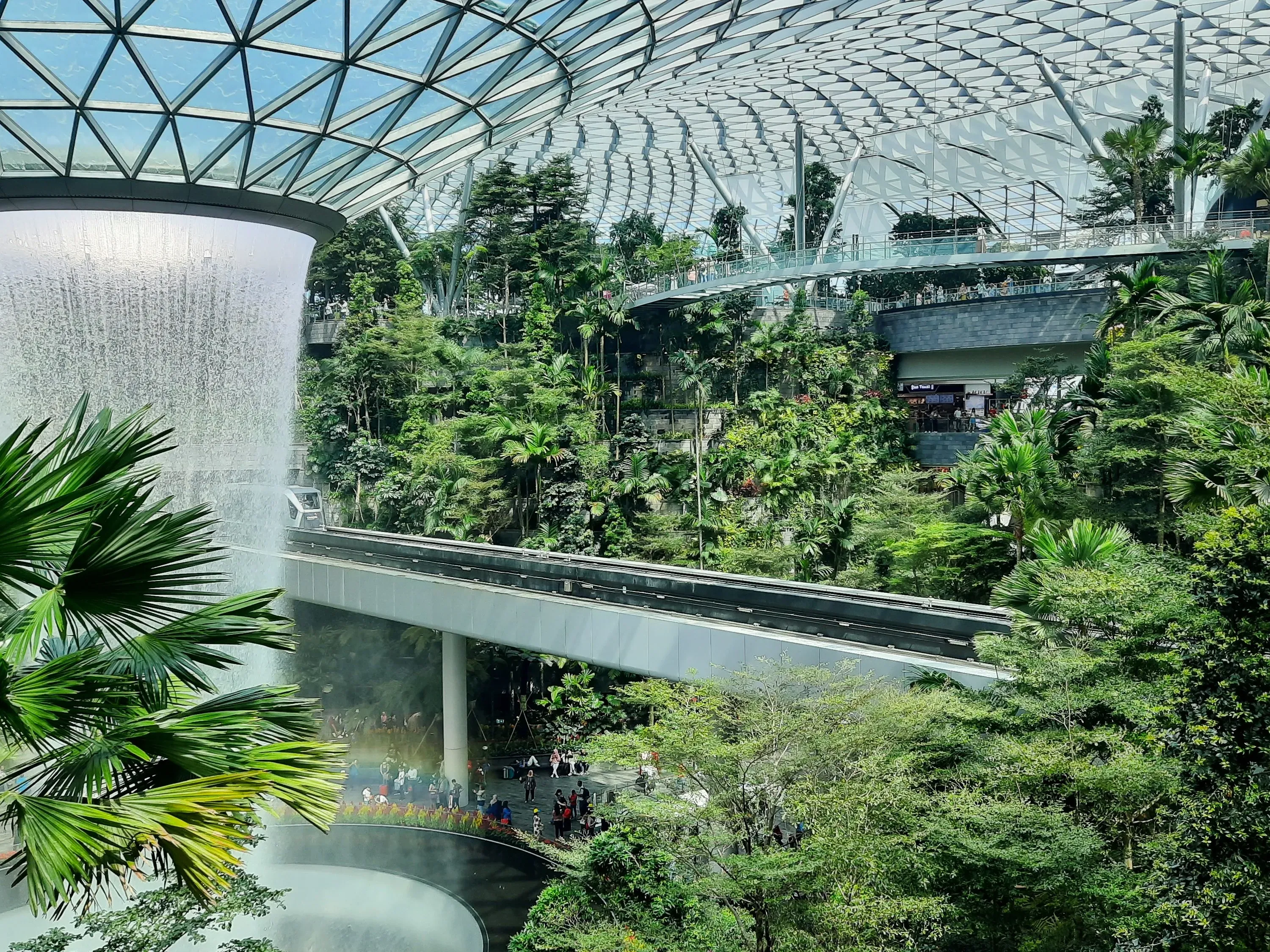 Wide view of the Rain Vortex waterfall inside Jewel Changi Airport, surrounded by indoor rainforest gardens and walkways.